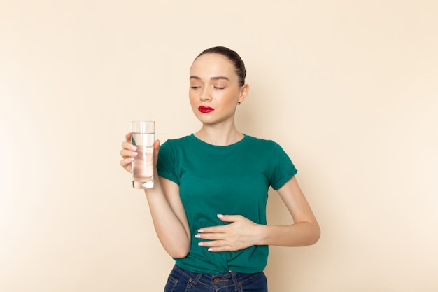 Vista frontal mujer joven en camisa verde oscuro y jeans con vaso de agua en beige