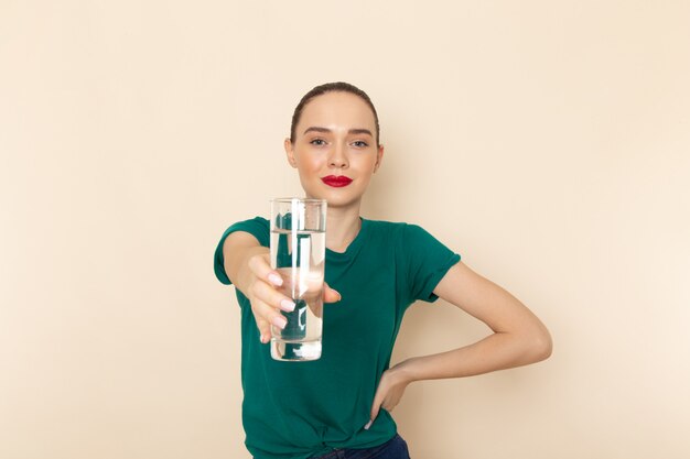 Vista frontal mujer joven en camisa verde oscuro y jeans con vaso de agua en beige