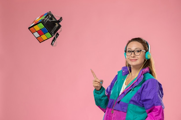 Vista frontal mujer joven en camiseta blanca abrigo de color con eraphones arrojando rubics cube bag sobre fondo rosa