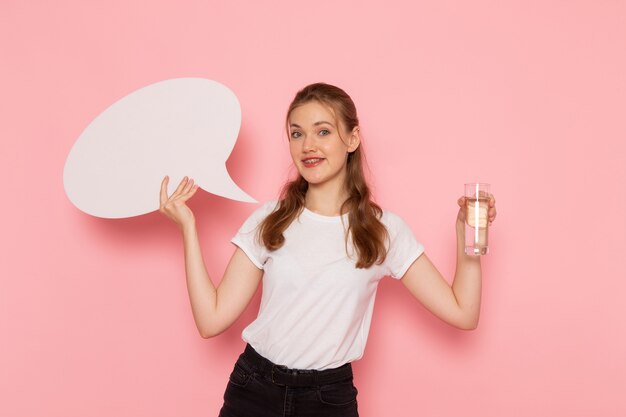 Vista frontal de la mujer joven en camiseta blanca con cartel blanco y vaso de agua en la pared rosa