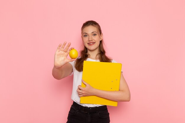 Vista frontal de la mujer joven en camiseta blanca con limón fresco con archivos sonriendo en la pared rosa