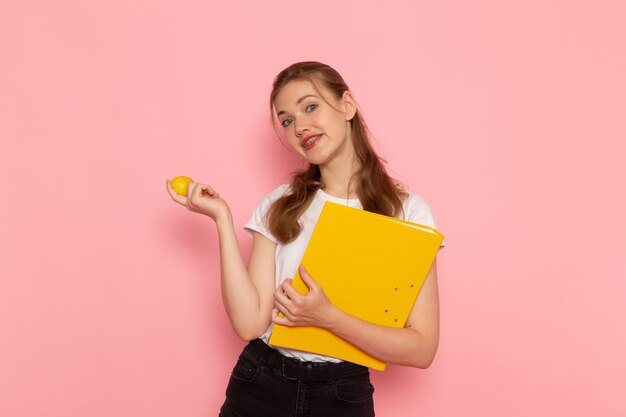 Vista frontal de la mujer joven en camiseta blanca con limón fresco con archivos sonriendo en la pared rosa