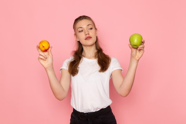 Vista frontal de la mujer joven en camiseta blanca con manzana verde fresca con melocotón en la pared de color rosa claro