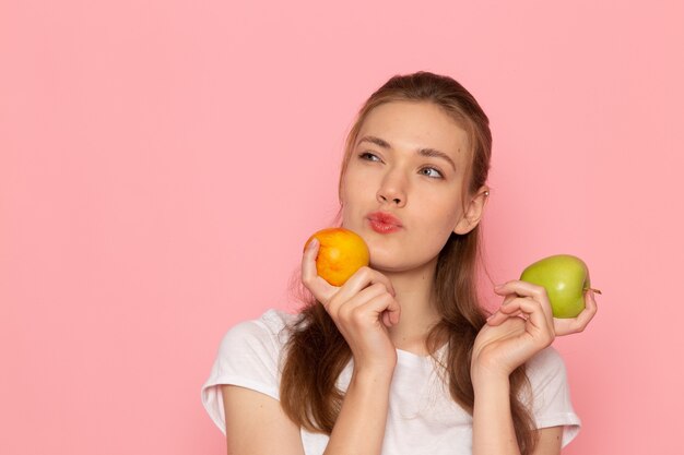 Vista frontal de la mujer joven en camiseta blanca con manzana verde fresca con melocotón pensando en la pared de color rosa claro