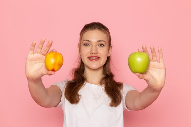 Vista frontal de la mujer joven en camiseta blanca con manzana verde fresca con melocotón sonriendo en la pared rosa claro