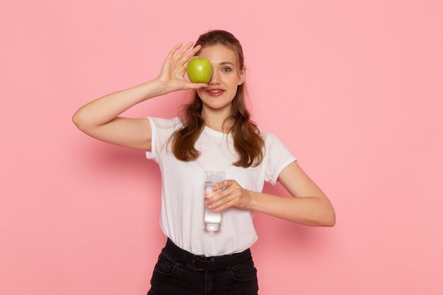 Vista frontal de la mujer joven en camiseta blanca con manzana verde fresca y vaso de agua en la pared rosa