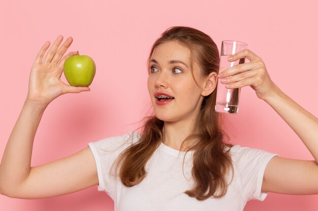 Vista frontal de la mujer joven en camiseta blanca con manzana verde fresca y vaso de agua en la pared rosa