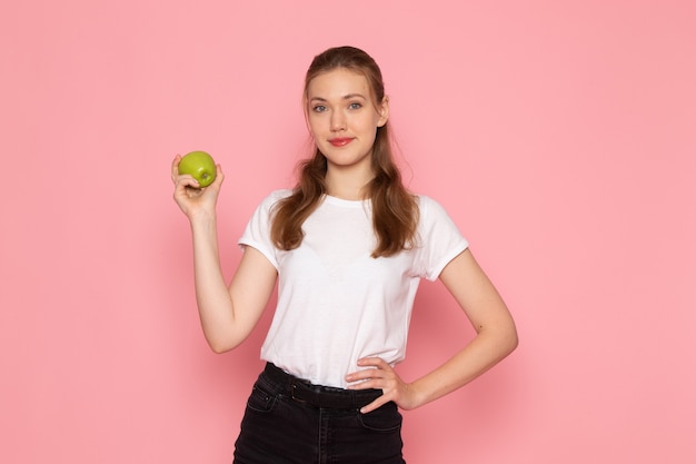 Vista frontal de la mujer joven en camiseta blanca con manzana verde en la pared rosa