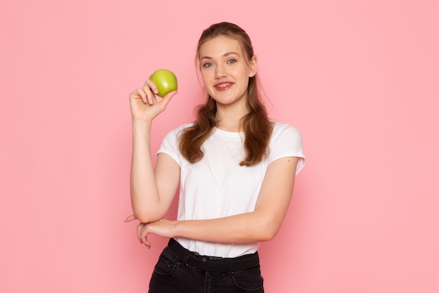 Vista frontal de la mujer joven en camiseta blanca con manzana verde sonriendo en la pared rosa