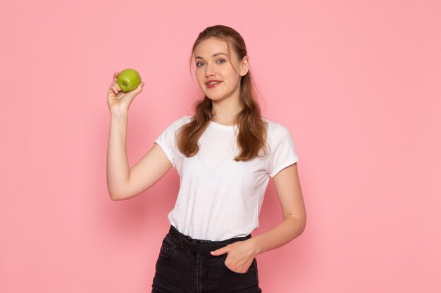 Vista frontal de la mujer joven en camiseta blanca con manzana verde sonriendo en la pared rosa
