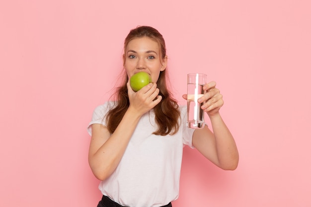 Vista frontal de la mujer joven en camiseta blanca con manzana verde y vaso de agua en la pared rosa