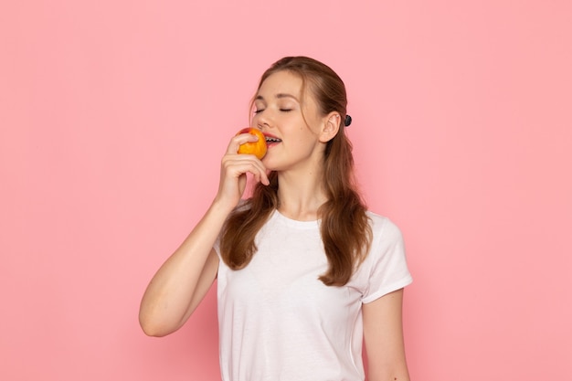 Vista frontal de la mujer joven en camiseta blanca con melocotón fresco comiéndolo en la pared de color rosa claro