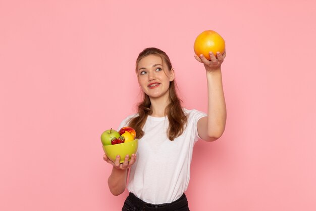 Vista frontal de la mujer joven en camiseta blanca con plato con frutas frescas y pomelo con sonrisa en la pared rosa