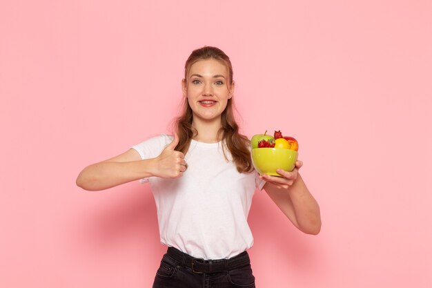 Vista frontal de la mujer joven en camiseta blanca con plato con frutas frescas sonriendo en la pared rosa