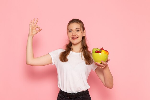 Vista frontal de la mujer joven en camiseta blanca con plato con frutas frescas sonriendo en la pared rosa
