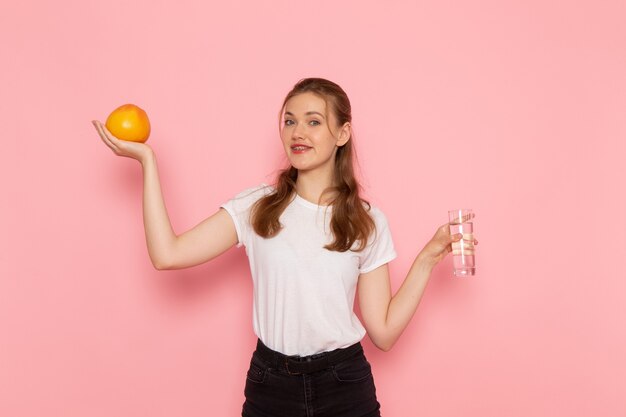 Vista frontal de la mujer joven en camiseta blanca con pomelo fresco y vaso de agua en la pared rosa