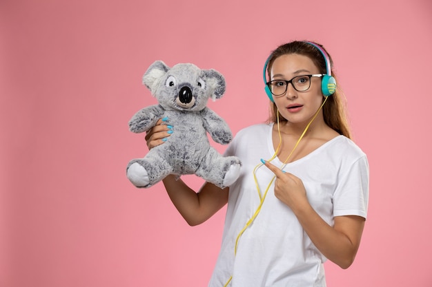 Vista frontal mujer joven en camiseta blanca solo escuchando música a través de auriculares y sosteniendo un lindo juguete en el escritorio rosa