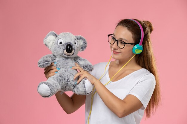 Vista frontal mujer joven en camiseta blanca solo escuchando música a través de auriculares y sosteniendo un lindo juguete en el fondo rosa