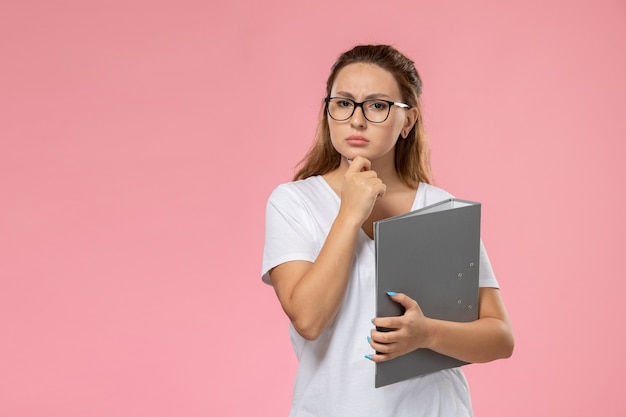 Foto gratuita vista frontal mujer joven en camiseta blanca sosteniendo documento gris con expresión de pensamiento sobre fondo rosa