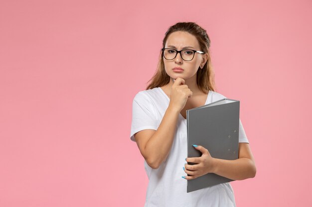 Vista frontal mujer joven en camiseta blanca sosteniendo documento gris con expresión de pensamiento sobre fondo rosa