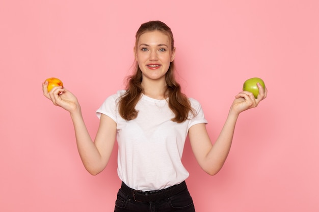 Vista frontal de la mujer joven en camiseta blanca sosteniendo una manzana verde fresca y sonriendo en la pared de color rosa claro