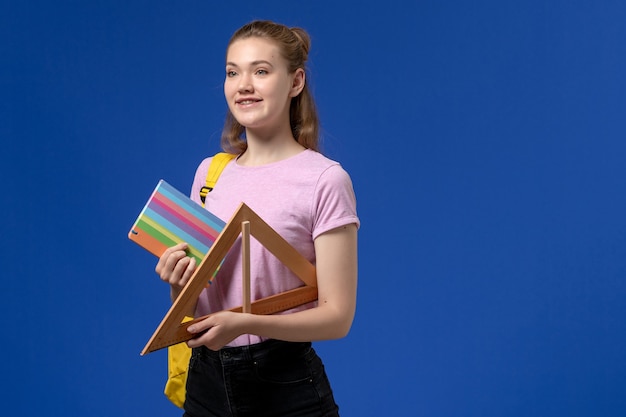 Vista frontal de la mujer joven en camiseta rosa con figura de triángulo de madera y cuaderno en la pared azul