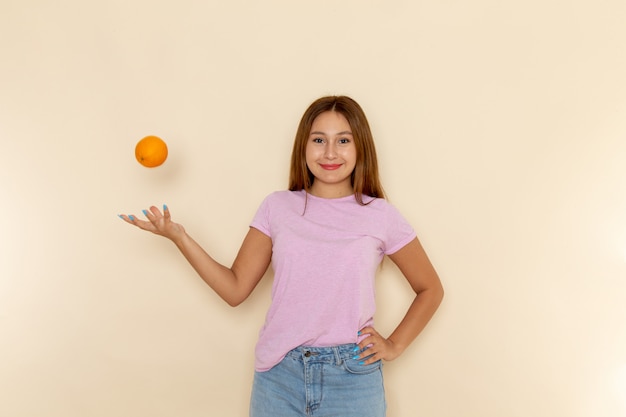 Vista frontal mujer joven en camiseta rosa y jeans azul sosteniendo naranja enfocándose con ella y sonriendo