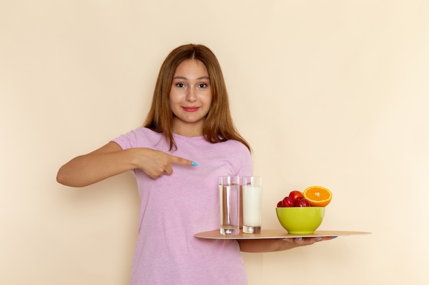 Vista frontal mujer joven en camiseta rosa y jeans con bandeja de frutas, leche y agua en gris