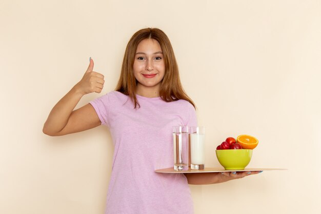 Vista frontal mujer joven en camiseta rosa y jeans con bandeja de frutas, leche y agua en gris