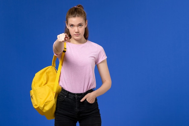 Vista frontal de la mujer joven en camiseta rosa con mochila amarilla apuntando hacia la pared azul claro