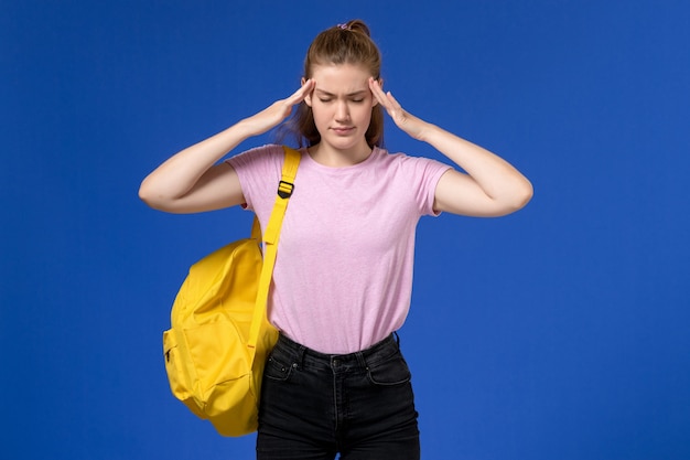 Vista frontal de la mujer joven en camiseta rosa con mochila amarilla con dolor de cabeza en la pared azul