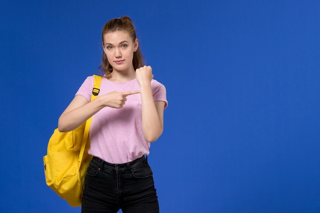 Vista frontal de la mujer joven en camiseta rosa con mochila amarilla mostrando su muñeca en la pared azul claro
