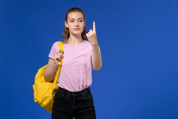 Vista frontal de la mujer joven en camiseta rosa con mochila amarilla en la pared azul claro