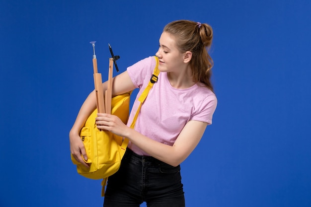 Vista frontal de la mujer joven en camiseta rosa con mochila amarilla en la pared azul