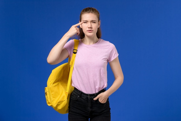 Vista frontal de la mujer joven en camiseta rosa con mochila amarilla pensando en la pared azul