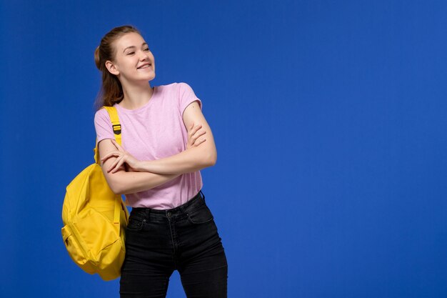 Vista frontal de la mujer joven en camiseta rosa con mochila amarilla posando y riendo en la pared azul