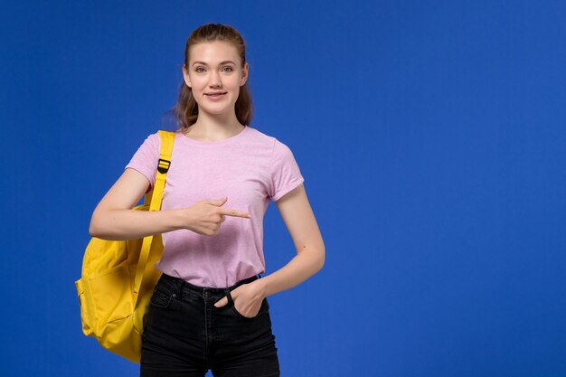 Vista frontal de la mujer joven en camiseta rosa con mochila amarilla posando con una sonrisa en la pared azul claro