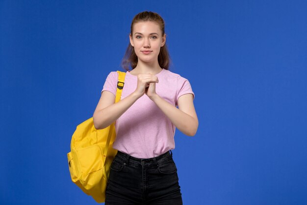 Vista frontal de la mujer joven en camiseta rosa con mochila amarilla sonriendo levemente en la pared azul