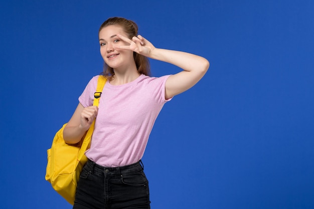 Vista frontal de la mujer joven en camiseta rosa con mochila amarilla sonriendo en la pared azul claro