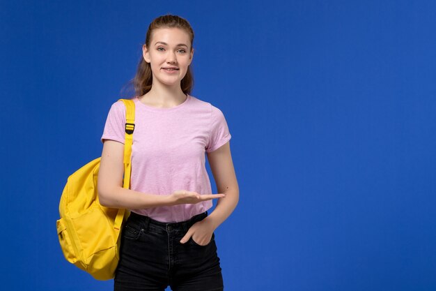 Vista frontal de la mujer joven en camiseta rosa con mochila amarilla sonriendo en la pared azul claro