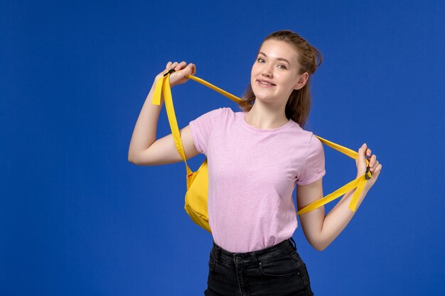 Vista frontal de la mujer joven en camiseta rosa con mochila amarilla sonriendo en la pared azul