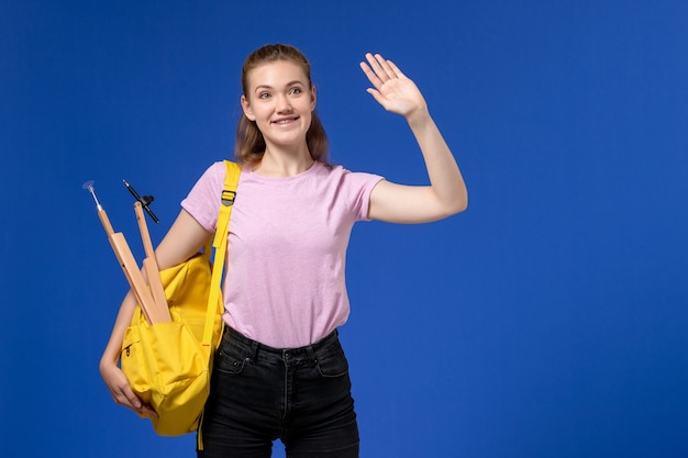 Vista frontal de la mujer joven en camiseta rosa con mochila amarilla sonriendo en la pared azul