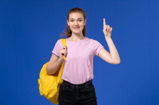 Vista frontal de la mujer joven en camiseta rosa con mochila amarilla sonriendo y posando en la pared azul