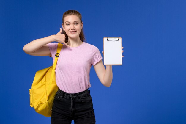 Vista frontal de la mujer joven en camiseta rosa con mochila amarilla sonriendo sosteniendo el bloc de notas en la pared azul