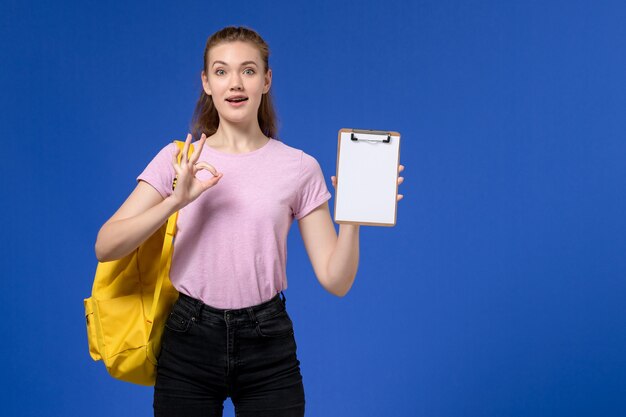 Vista frontal de la mujer joven en camiseta rosa con mochila amarilla y sosteniendo el bloc de notas posando en la pared azul