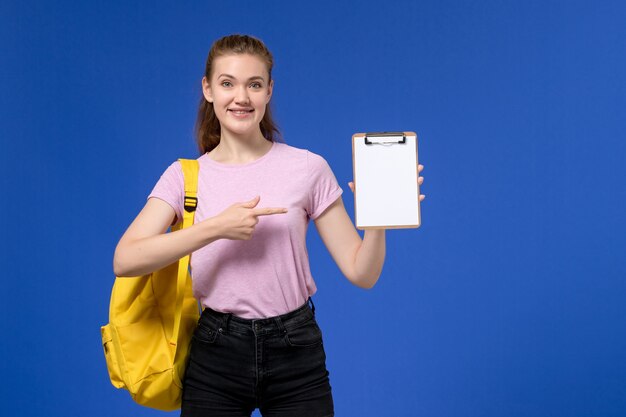 Vista frontal de la mujer joven en camiseta rosa con mochila amarilla y sosteniendo el bloc de notas sonriendo en la pared azul