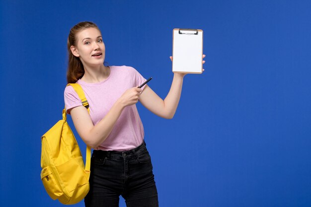 Vista frontal de la mujer joven en camiseta rosa con mochila amarilla y sosteniendo el bloc de notas