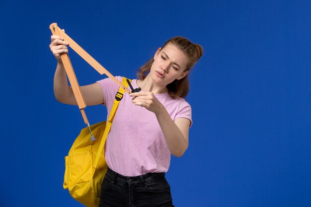 Vista frontal de la mujer joven en camiseta rosa con mochila amarilla sosteniendo una figura de madera en la pared azul