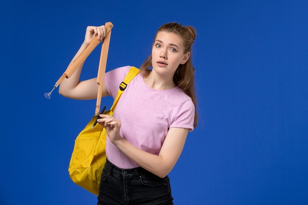 Vista frontal de la mujer joven en camiseta rosa con mochila amarilla sosteniendo una figura de madera en la pared azul