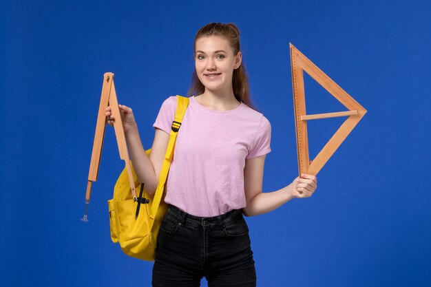 Vista frontal de la mujer joven en camiseta rosa con mochila amarilla sosteniendo figuras de madera en la pared azul